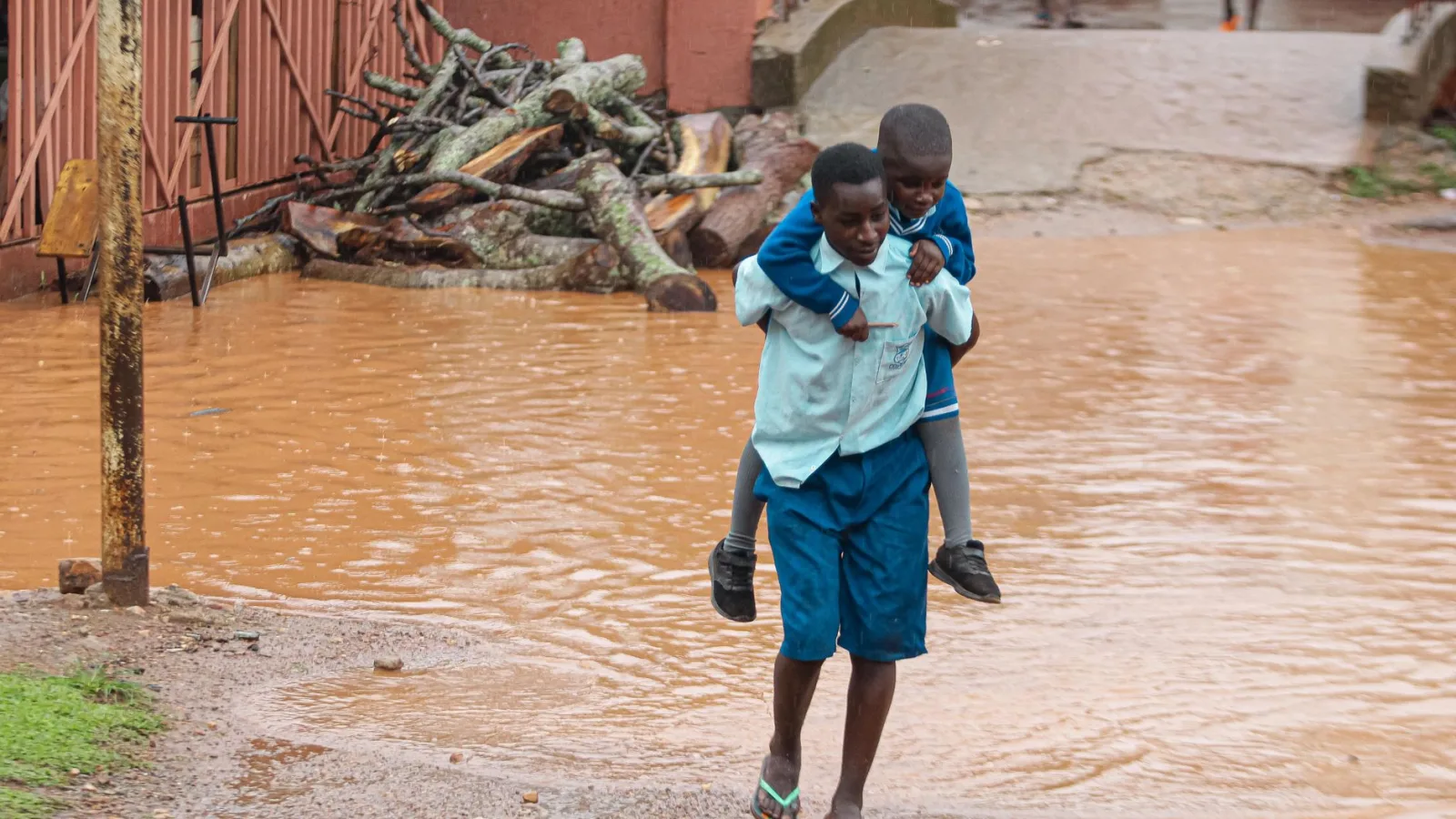 A young boy piggybacks his friend across a flooded muddy street in a village setting.