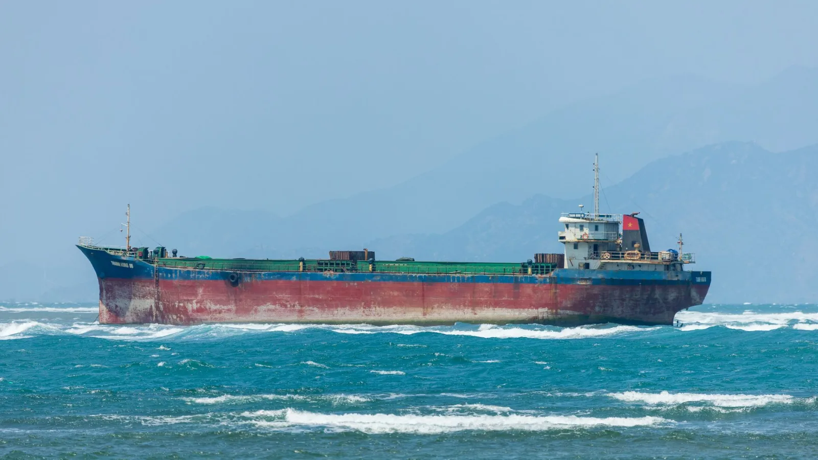 A vast cargo ship sails through rough seas with a mountainous backdrop.