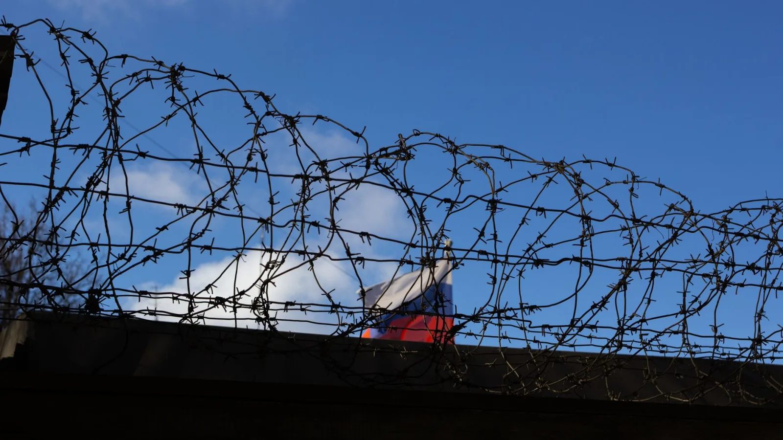 Silhouette of barbed wire with a flag in the background against a clear blue sky.