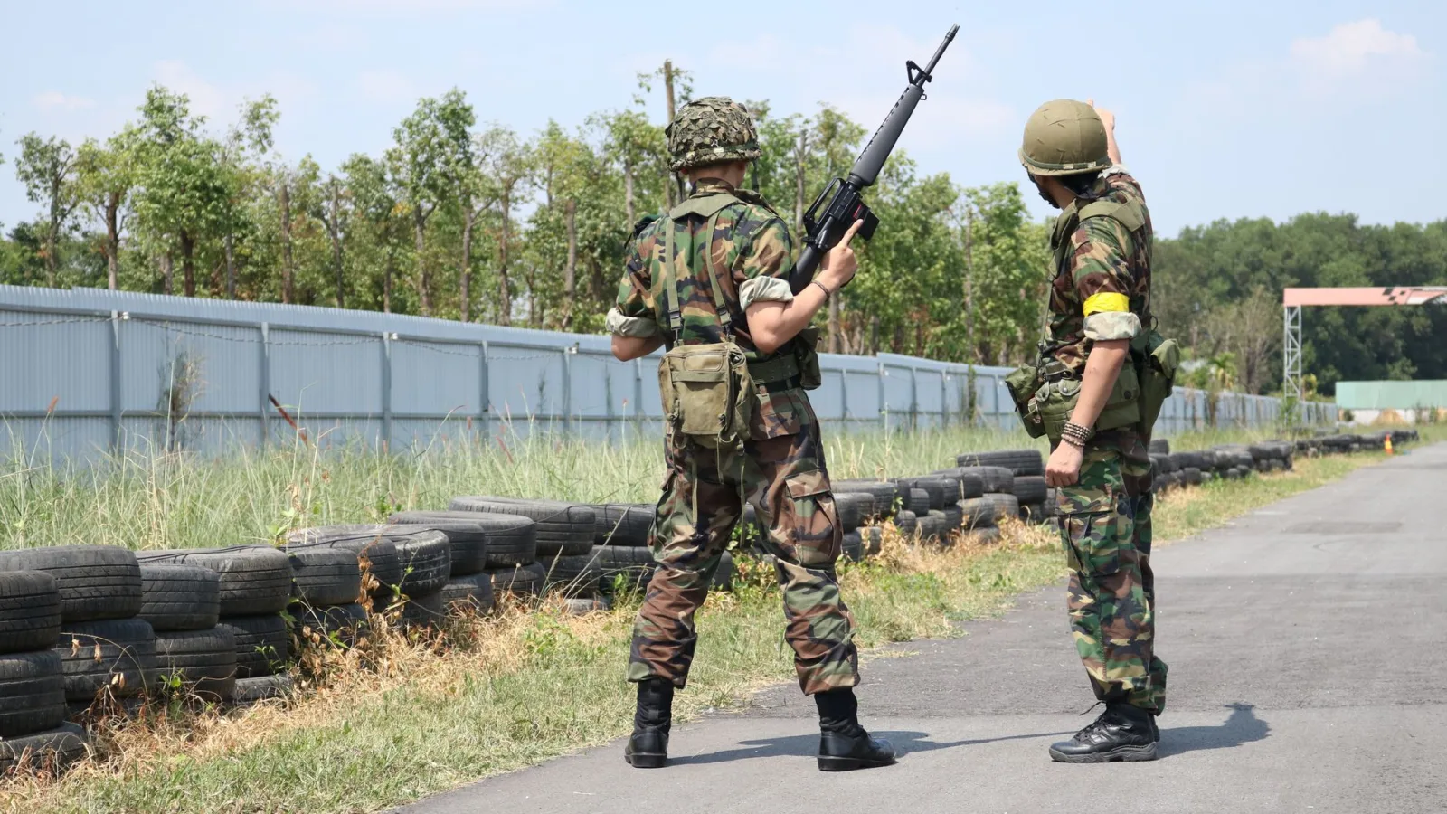 Two soldiers in camouflage uniforms with weapons stand outside on a training field.