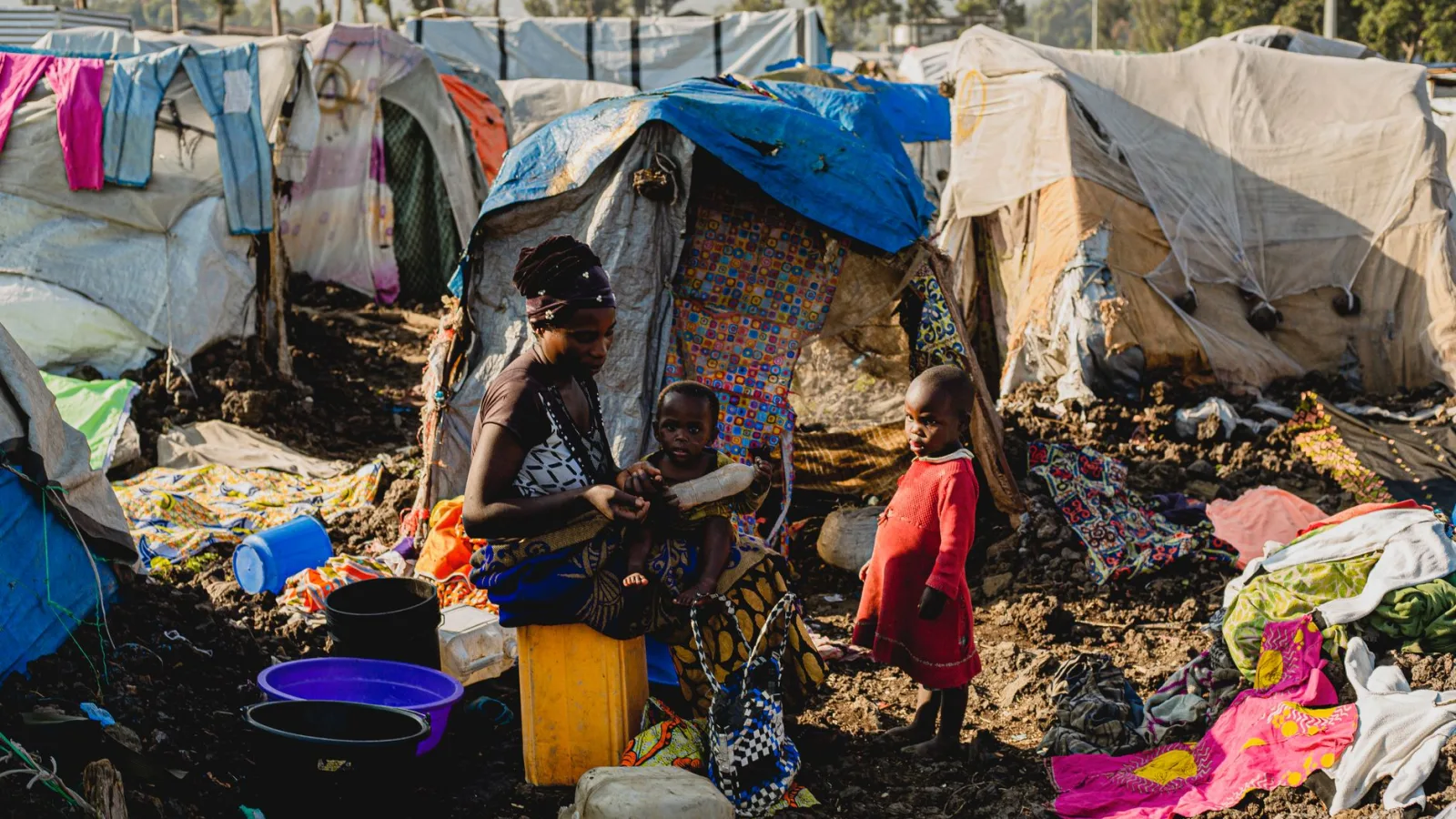 Black woman with children in a makeshift camp setting amidst tents and scattered items.