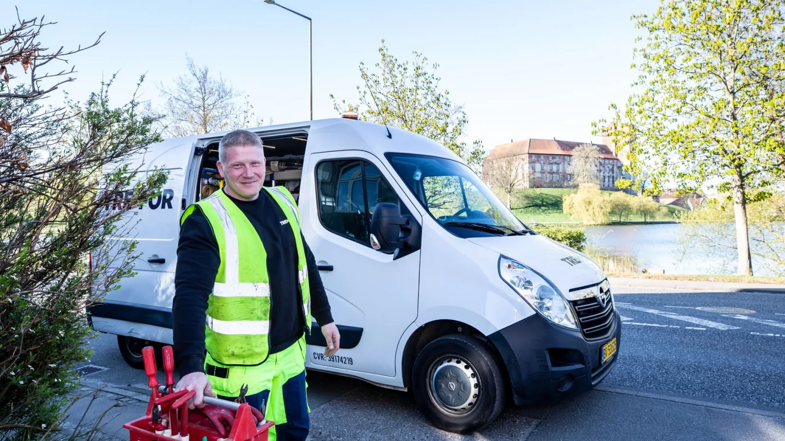 Smiling worker in reflective vest stands by a van holding tools.