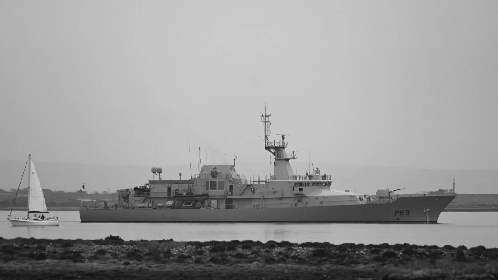 Black and white image of a naval patrol ship alongside a sailboat on calm waters.