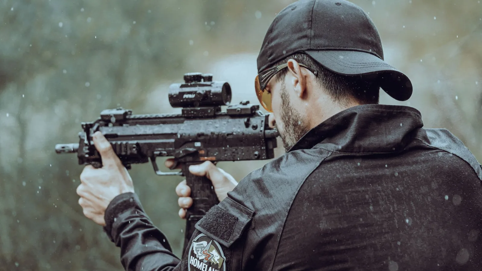 Man aiming with tactical gear and firearm in an outdoor setting during rain.